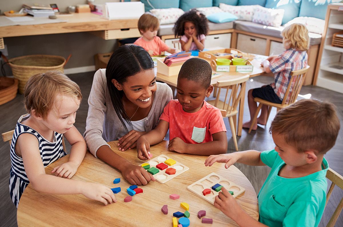 A teacher smiling as she works through a shape puzzle with three young children at a round classroom table