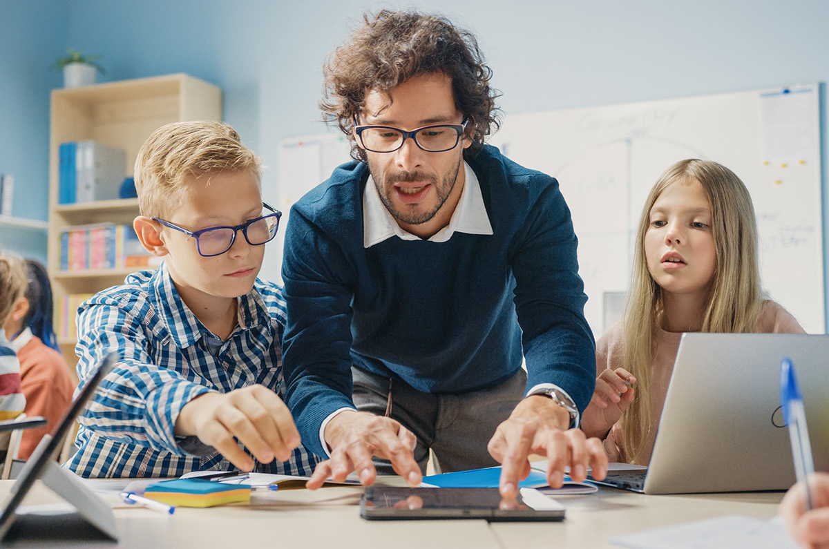 A teacher guiding two students through a lesson on a tablet in a bright classroom