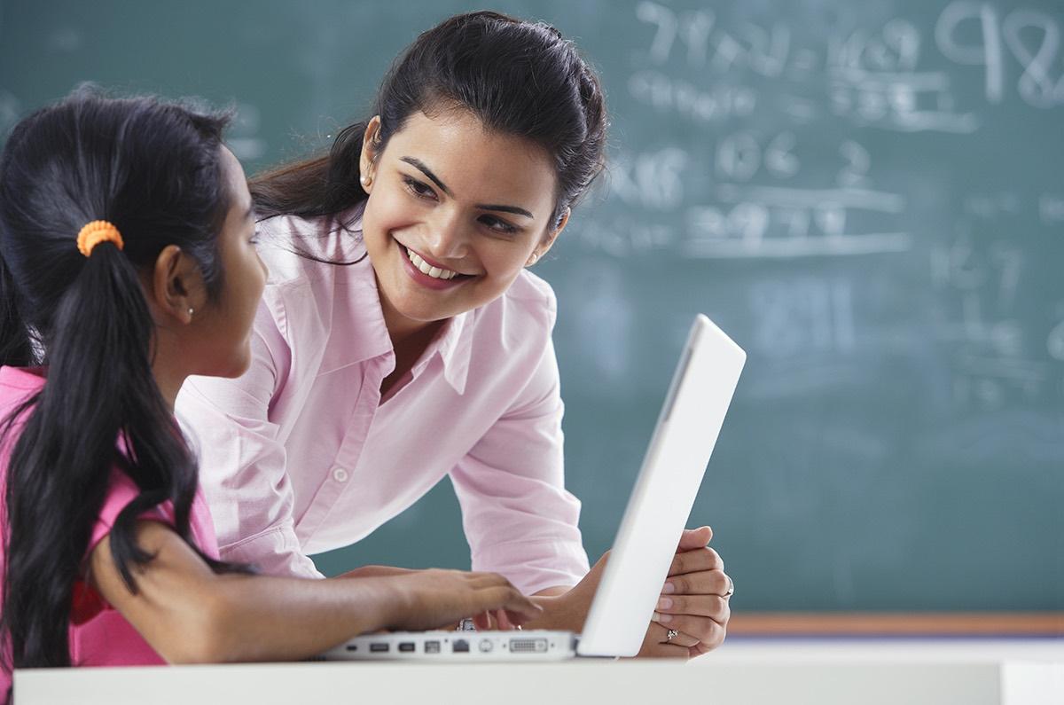 A teacher leaning in to help a girl working on a laptop in front of a chalkboard